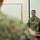 A New Zealand Army soldier, identified by 'N.Z. ARMY' text on their uniform, is addressing a group of military personnel in what appears to be a classroom or briefing room setting. The speaker is gesturing with their hands while speaking to seated audience members whose backs are visible in the foreground. The setting suggests a formal military training or instructional session.