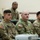 A group of U.S. Army soldiers in OCP (Operational Camouflage Pattern) uniforms are seated at desks or tables, appearing to attend a briefing, training session, or classroom instruction. American flag patches are visible on their sleeves, and at least one name tape and 'U.S. ARMY' tape are legible. A projector or electronic device is visible in the foreground.