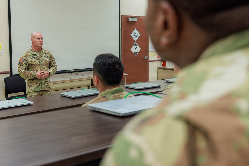 A U.S. Army soldier, appearing to be a senior non-commissioned officer or officer, stands at the front of a classroom briefing or instructing a small group of soldiers seated at a conference table. The setting appears to be a military training or administrative facility, with laptops placed in front of each seated soldier. The instructor has his hands clasped and is addressing the group in front of a whiteboard.