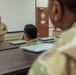 A U.S. Army soldier, appearing to be a senior non-commissioned officer or officer, stands at the front of a classroom briefing or instructing a small group of soldiers seated at a conference table. The setting appears to be a military training or administrative facility, with laptops placed in front of each seated soldier. The instructor has his hands clasped and is addressing the group in front of a whiteboard.