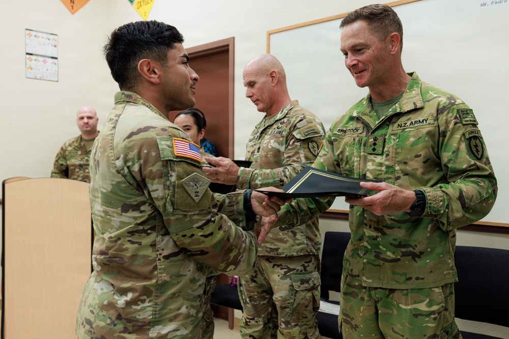 A military ceremony is taking place in what appears to be a classroom or briefing room, where a document or award folder is being exchanged between a U.S. Army soldier and a New Zealand Army officer identified by his 'N.Z. ARMY' name tape and 'SHYSTOCK' surname tape. A third U.S. Army soldier stands in the background observing the exchange. The setting and formal nature of the handoff suggest an award, commendation, or certificate presentation ceremony.