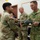 A military ceremony is taking place in what appears to be a classroom or briefing room, where a document or award folder is being exchanged between a U.S. Army soldier and a New Zealand Army officer identified by his 'N.Z. ARMY' name tape and 'SHYSTOCK' surname tape. A third U.S. Army soldier stands in the background observing the exchange. The setting and formal nature of the handoff suggest an award, commendation, or certificate presentation ceremony.