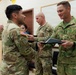 A military ceremony is taking place in what appears to be a classroom or briefing room, where a document or award folder is being exchanged between a U.S. Army soldier and a New Zealand Army officer identified by his 'N.Z. ARMY' name tape and 'SHYSTOCK' surname tape. A third U.S. Army soldier stands in the background observing the exchange. The setting and formal nature of the handoff suggest an award, commendation, or certificate presentation ceremony.