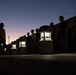 A low-light dusk photograph showing a uniformed authority figure facing a line of individuals standing at attention outdoors. The scene is dramatically silhouetted against a twilight sky, with a row of illuminated stone pathway lanterns providing the primary light source. The composition suggests a formal inspection, formation, or orientation event.