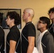 Five young men are standing in a line facing the same direction in profile view, inside a room with a yellow wall. Most are wearing matching black polo shirts with white trim detailing, suggesting a uniform or team affiliation. A framed photograph or artwork is visible on the wall behind them.