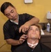 A barber is cutting the hair of a male client in a barbershop setting. The barber, dressed in a black uniform, is using electric clippers on the side of the client's head while the client sits reclined with a black cape and white neck strip. The setting appears to be a professional barbershop with tools and products visible on a counter in the background.