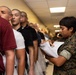 A U.S. Marines service member in digital camouflage uniform reviews a stack of papers/documents while standing beside a line of civilians in a corridor. The civilians appear to be recruits or inductees, several wearing minimal or plain clothing, suggesting a processing or in-processing event. The setting appears to be an indoor military facility hallway.
