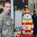 A U.S. Air Force servicemember named Johnston is speaking at what appears to be a fire safety awareness event inside a fire station. Behind him is a Sparky the Fire Dog mascot costume/figure dressed in firefighter gear, and a civilian man in a dark shirt stands in the background. A document or sign is visible on a stand beside the speaker.