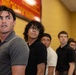A line of young men standing along a yellow wall, appearing to wait for something. The foreground subject, wearing a gray polo shirt, is looking upward with an alert or anxious expression. The scene suggests a queue or lineup in an institutional or public setting.