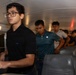 A group of young men are standing in the aisle of what appears to be a school bus or charter bus. The individuals are lined up single-file along the center aisle, with bus seats visible on both sides. The interior lighting is dim, with small overhead lights illuminating the space.