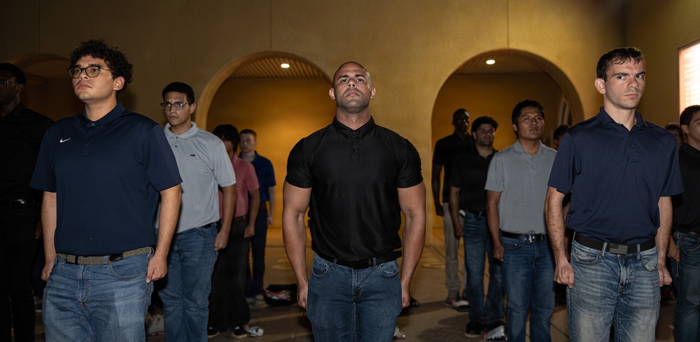 A group of men stand in a formal, disciplined formation inside a building with arched architectural features and warm ambient lighting. The individuals appear to be at attention, with serious expressions and rigid postures suggesting a structured or ceremonial context. The scene resembles an orientation, pledge ceremony, or organized group gathering.