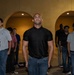 A group of men stand in a formal, disciplined formation inside a building with arched architectural features and warm ambient lighting. The individuals appear to be at attention, with serious expressions and rigid postures suggesting a structured or ceremonial context. The scene resembles an orientation, pledge ceremony, or organized group gathering.