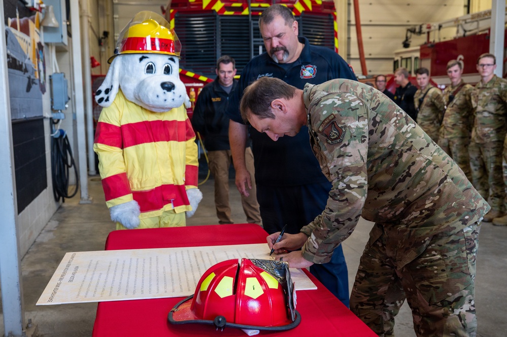 A military officer in OCP (Operational Camouflage Pattern) uniform is signing an official document at a table inside a fire station bay. The ceremony includes civilian fire department personnel, military personnel in the background, a red firefighter helmet on the table, and a Dalmatian fire safety mascot costume (resembling 'Sparky the Fire Dog'). This appears to be a formal signing ceremony between a military installation and a fire department.