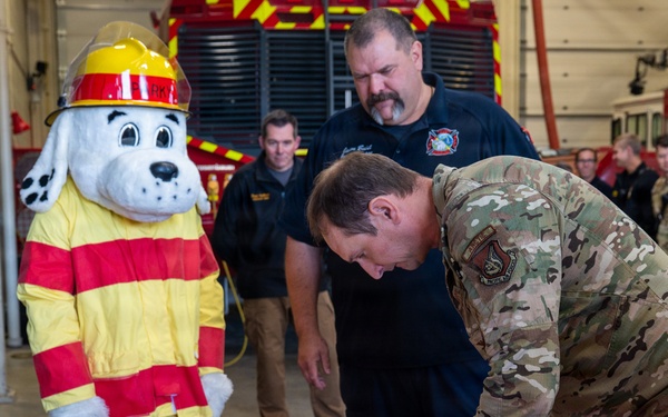 A military officer in OCP (Operational Camouflage Pattern) uniform is signing an official document at a table inside a fire station bay. The ceremony includes civilian fire department personnel, military personnel in the background, a red firefighter helmet on the table, and a Dalmatian fire safety mascot costume (resembling 'Sparky the Fire Dog'). This appears to be a formal signing ceremony between a military installation and a fire department.