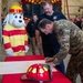 A military officer in OCP (Operational Camouflage Pattern) uniform is signing an official document at a table inside a fire station bay. The ceremony includes civilian fire department personnel, military personnel in the background, a red firefighter helmet on the table, and a Dalmatian fire safety mascot costume (resembling 'Sparky the Fire Dog'). This appears to be a formal signing ceremony between a military installation and a fire department.