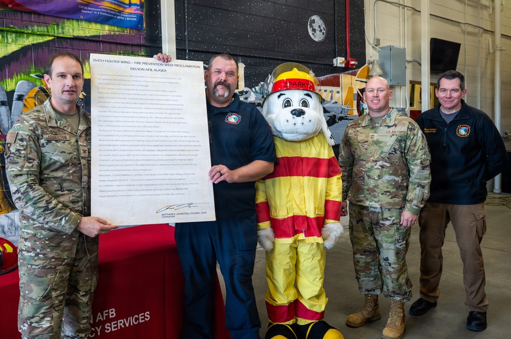 Five individuals pose for a group photo at Eielson AFB, Alaska, during a Fire Prevention Week Proclamation ceremony for the 354th Fighter Wing. The group includes military personnel in Army Combat Uniforms, a civilian firefighter, a fire safety mascot character named 'Sparky' the Dalmatian, and a civilian official. One person holds a large framed proclamation document titled '354th Fighter Wing – Fire Prevention Week Proclamation, Eielson AFB, Alaska.'