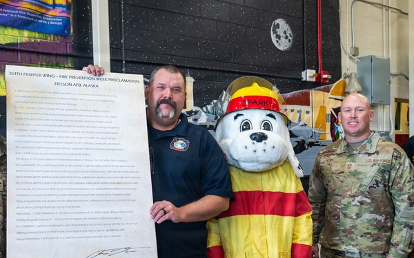 Five individuals pose for a group photo at Eielson AFB, Alaska, during a Fire Prevention Week Proclamation ceremony for the 354th Fighter Wing. The group includes military personnel in Army Combat Uniforms, a civilian firefighter, a fire safety mascot character named 'Sparky' the Dalmatian, and a civilian official. One person holds a large framed proclamation document titled '354th Fighter Wing – Fire Prevention Week Proclamation, Eielson AFB, Alaska.'