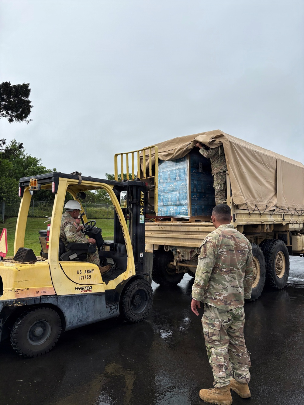 US Army personnel are loading or unloading pallets of bottled water onto a military cargo truck using a Hyster Fortis forklift (serial number 171769) marked 'US ARMY'. The operation is taking place on a wet surface, suggesting recent rainfall, with at least three soldiers visible in military camouflage (OCP pattern) uniforms. This appears to be a logistics or humanitarian relief operation involving water distribution.