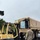 US Army personnel are loading or unloading pallets of bottled water onto a military cargo truck using a Hyster Fortis forklift (serial number 171769) marked 'US ARMY'. The operation is taking place on a wet surface, suggesting recent rainfall, with at least three soldiers visible in military camouflage (OCP pattern) uniforms. This appears to be a logistics or humanitarian relief operation involving water distribution.