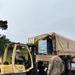 US Army personnel are loading or unloading pallets of bottled water onto a military cargo truck using a Hyster Fortis forklift (serial number 171769) marked 'US ARMY'. The operation is taking place on a wet surface, suggesting recent rainfall, with at least three soldiers visible in military camouflage (OCP pattern) uniforms. This appears to be a logistics or humanitarian relief operation involving water distribution.