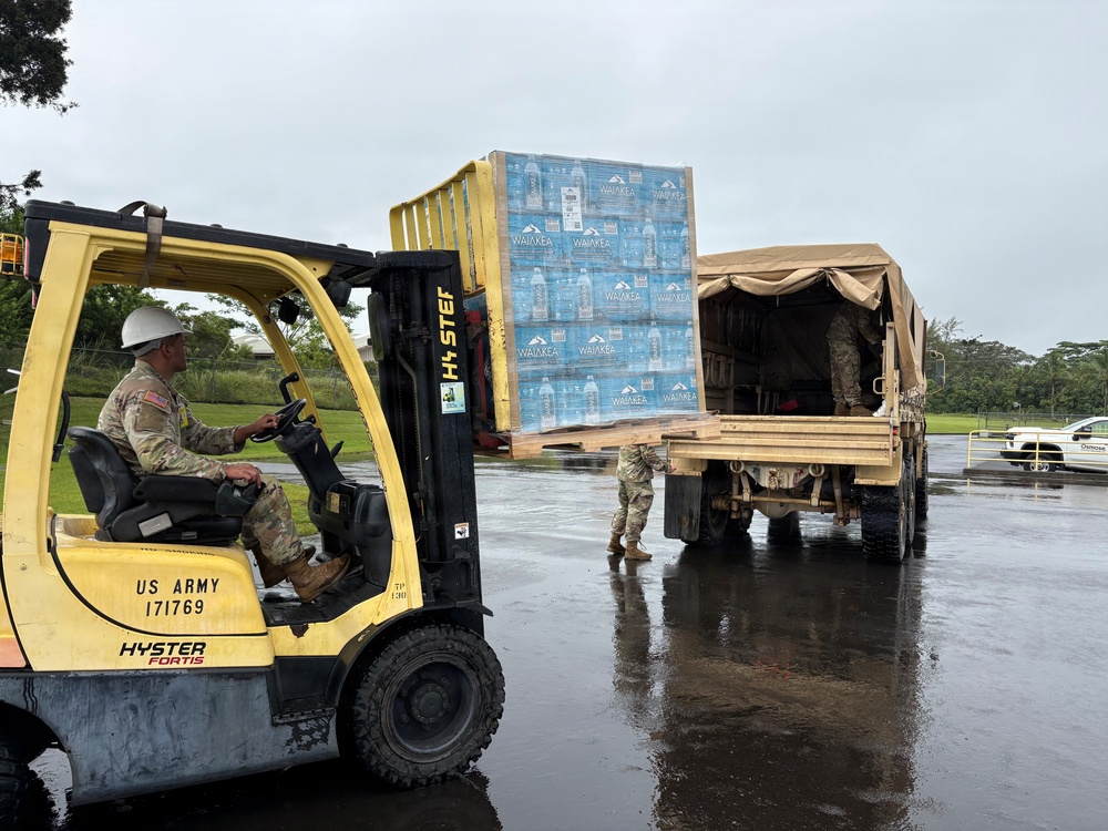 U.S. Army personnel are loading pallets of Waiākea bottled water onto a military cargo truck using a yellow Hyster Fortis forklift marked 'US ARMY 171769'. The operation appears to be a humanitarian or disaster relief supply distribution effort, taking place on a wet surface suggesting recent rainfall. At least three soldiers in camouflage uniforms are visible participating in the logistics operation.