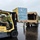 U.S. Army personnel are loading pallets of Waiākea bottled water onto a military cargo truck using a yellow Hyster Fortis forklift marked 'US ARMY 171769'. The operation appears to be a humanitarian or disaster relief supply distribution effort, taking place on a wet surface suggesting recent rainfall. At least three soldiers in camouflage uniforms are visible participating in the logistics operation.