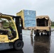 U.S. Army personnel are loading pallets of Waiākea bottled water onto a military cargo truck using a yellow Hyster Fortis forklift marked 'US ARMY 171769'. The operation appears to be a humanitarian or disaster relief supply distribution effort, taking place on a wet surface suggesting recent rainfall. At least three soldiers in camouflage uniforms are visible participating in the logistics operation.