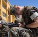 A U.S. Marine in MARPAT woodland digital camouflage uniform is focused on cleaning or inspecting a rifle, likely an M16 or M4 variant, outdoors in a sunny environment. Several other Marines in the same uniform are visible in the background performing similar tasks. The setting appears to be a military installation courtyard or parade deck adjacent to a yellow building.