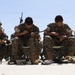 A group of military personnel in camouflage uniforms are seated on folding stools performing weapons maintenance on their rifles. The scene appears to be an outdoor field training or cleaning exercise conducted on a paved surface in sunny conditions. Multiple service members are visible in the background performing the same activity.