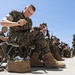 A group of U.S. Marines in MARPAT digital camouflage uniforms are seated outdoors on folding stools, performing weapons maintenance on their rifles. The foreground Marine is focused on cleaning or inspecting a rifle component, with a tan gear bag at his feet. The setting appears to be a military installation, likely a training base, with a yellow building visible in the background.