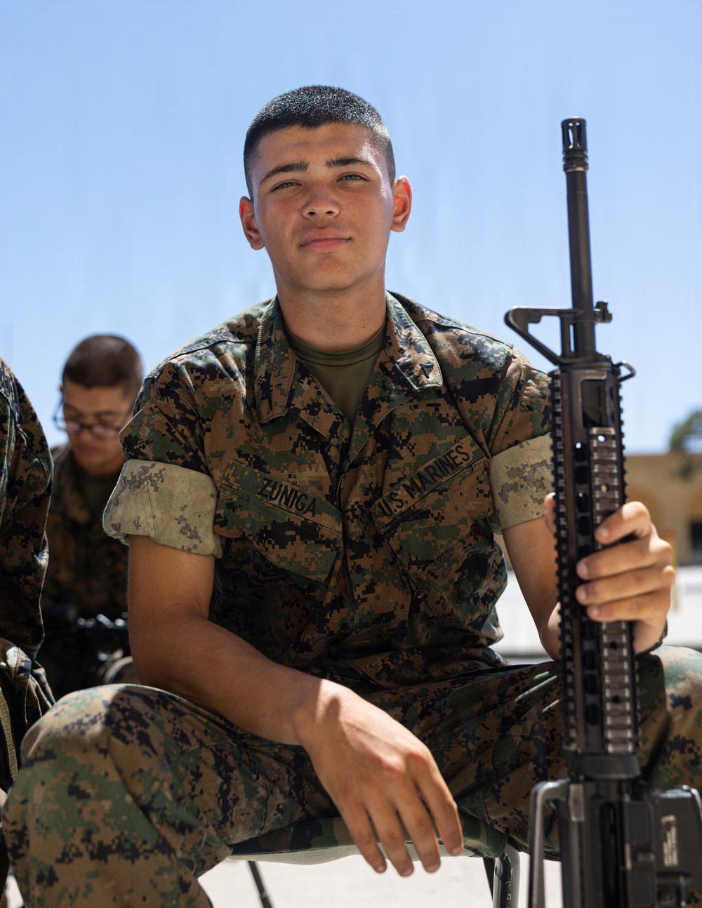 A young U.S. Marine wearing MARPAT digital camouflage utilities is seated outdoors holding a rifle, identified by the name tape 'ZUNIGA' and 'U.S. MARINES' tape on his uniform. He is smiling toward the camera in what appears to be a training or instructional setting. Other Marines in similar uniforms are visible in the background.