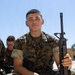 A young U.S. Marine wearing MARPAT digital camouflage utilities is seated outdoors holding a rifle, identified by the name tape 'ZUNIGA' and 'U.S. MARINES' tape on his uniform. He is smiling toward the camera in what appears to be a training or instructional setting. Other Marines in similar uniforms are visible in the background.