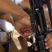 A close-up photograph showing hands manipulating what appears to be an M16 or AR-15 style rifle, specifically working on the upper receiver area. The person is wearing tan/khaki military-style clothing and appears to be performing maintenance or assembly on the firearm. Markings visible on the barrel include 'R14' and 'R16'.