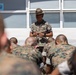 A U.S. Marine drill instructor wearing a campaign cover (drill sergeant hat) addresses a group of recruits or junior Marines seated in front of him. The instructor is standing and appears to be speaking or briefing the assembled personnel, who are wearing MARPAT camouflage uniforms. The setting appears to be an outdoor area adjacent to a building.