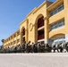 A large group of military personnel in camouflage uniforms are seated on folding chairs in a long row outside a large yellow/ochre-colored institutional building. They appear to be engaged in individual tasks, possibly cleaning weapons or studying materials. The setting includes palm trees and a clear blue sky, suggesting a warm, sunny climate consistent with a Southern California military installation.