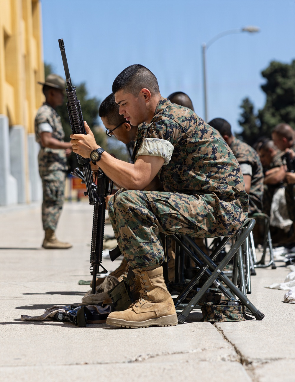 A group of military personnel in MARPAT digital camouflage uniforms are seated on folding stools outdoors, engaged in rifle maintenance or inspection. The foreground subject is closely examining an M16-style rifle while seated, with cleaning equipment visible on the ground. The setting appears to be a military base or training facility during daytime.