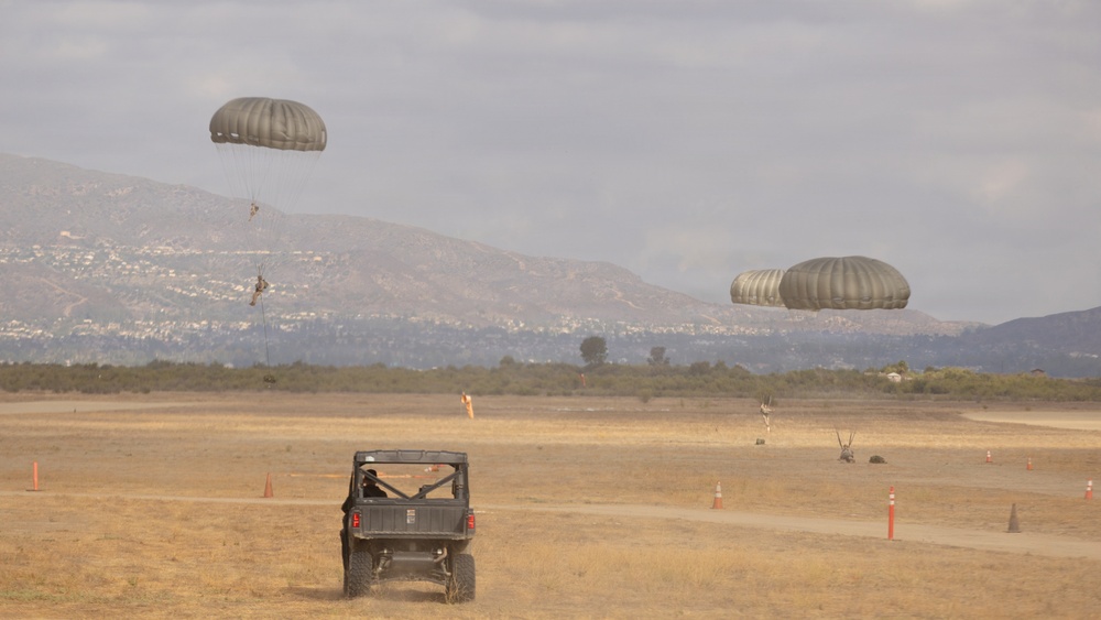 A military parachute training exercise is underway on a dry, open drop zone with two parachutists descending under olive-drab round canopies. A utility terrain vehicle (UTV) is positioned on the field, likely serving as a safety or recovery vehicle. Orange traffic cones mark the landing zone boundaries, with mountainous terrain and a hazy sky visible in the background.