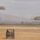 A military parachute training exercise is underway on a dry, open drop zone with two parachutists descending under olive-drab round canopies. A utility terrain vehicle (UTV) is positioned on the field, likely serving as a safety or recovery vehicle. Orange traffic cones mark the landing zone boundaries, with mountainous terrain and a hazy sky visible in the background.