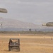 A military parachute training exercise is underway on a dry, open drop zone with two parachutists descending under olive-drab round canopies. A utility terrain vehicle (UTV) is positioned on the field, likely serving as a safety or recovery vehicle. Orange traffic cones mark the landing zone boundaries, with mountainous terrain and a hazy sky visible in the background.