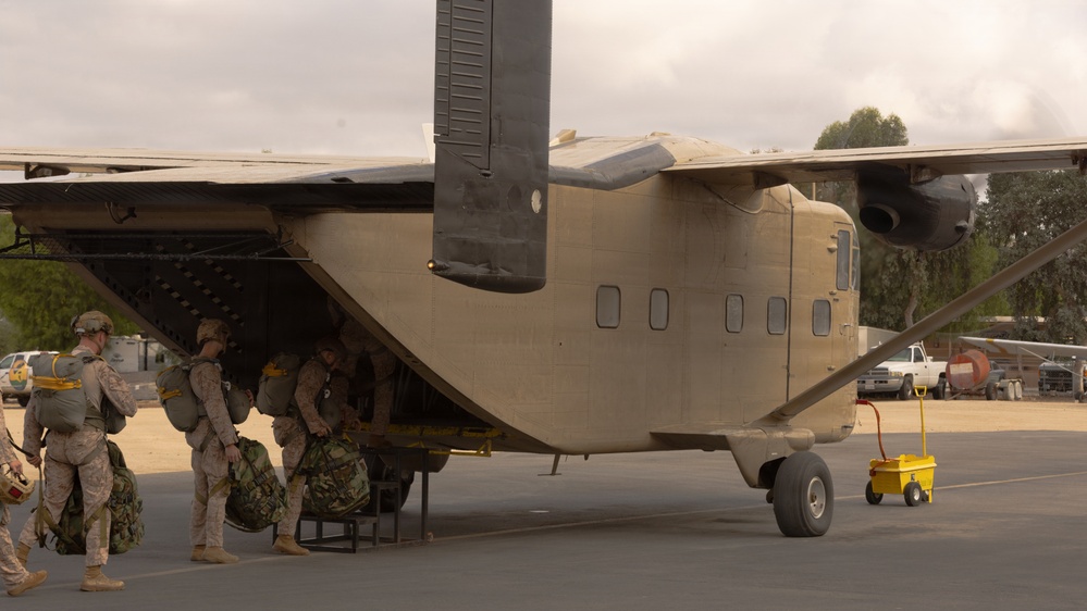 Military personnel in desert camouflage uniforms are loading gear and equipment into the rear ramp of a sand/tan-colored fixed-wing military transport aircraft. The aircraft appears to be a C-23 Sherpa or similar short takeoff and landing (STOL) transport, identifiable by its high-wing configuration and rear-loading ramp. The scene takes place on a tarmac at what appears to be a military airfield with trees and vehicles visible in the background.