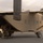 Military personnel in desert camouflage uniforms are loading gear and equipment into the rear ramp of a sand/tan-colored fixed-wing military transport aircraft. The aircraft appears to be a C-23 Sherpa or similar short takeoff and landing (STOL) transport, identifiable by its high-wing configuration and rear-loading ramp. The scene takes place on a tarmac at what appears to be a military airfield with trees and vehicles visible in the background.