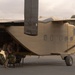 Military personnel in desert camouflage uniforms are loading gear and equipment into the rear ramp of a sand/tan-colored fixed-wing military transport aircraft. The aircraft appears to be a C-23 Sherpa or similar short takeoff and landing (STOL) transport, identifiable by its high-wing configuration and rear-loading ramp. The scene takes place on a tarmac at what appears to be a military airfield with trees and vehicles visible in the background.