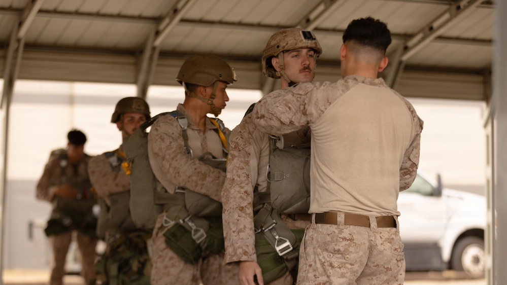 Military personnel in desert camouflage uniforms are conducting pre-jump equipment checks under a covered structure. One service member wearing a combat helmet and parachute harness is being inspected by another who appears to be checking harness fittings and connections. Additional personnel with parachute equipment are visible in the background, suggesting a pre-airborne operation preparation sequence.