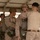 Military personnel in desert camouflage uniforms are conducting pre-jump equipment checks under a covered structure. One service member wearing a combat helmet and parachute harness is being inspected by another who appears to be checking harness fittings and connections. Additional personnel with parachute equipment are visible in the background, suggesting a pre-airborne operation preparation sequence.