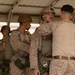 Military personnel in desert camouflage uniforms are conducting pre-jump equipment checks under a covered structure. One service member wearing a combat helmet and parachute harness is being inspected by another who appears to be checking harness fittings and connections. Additional personnel with parachute equipment are visible in the background, suggesting a pre-airborne operation preparation sequence.