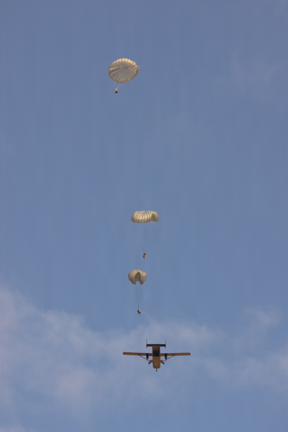 A military static-line parachute operation is captured in progress, showing three paratroopers at different descent stages above a twin-engine turboprop aircraft. The parachutes are tan/olive-colored round canopies consistent with military-issue static-line parachutes. The aircraft below appears to be a C-23 Sherpa or similar twin-boom military transport, viewed from below as it continues its drop run.