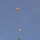 A military static-line parachute operation is captured in progress, showing three paratroopers at different descent stages above a twin-engine turboprop aircraft. The parachutes are tan/olive-colored round canopies consistent with military-issue static-line parachutes. The aircraft below appears to be a C-23 Sherpa or similar twin-boom military transport, viewed from below as it continues its drop run.