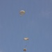 A military static-line parachute operation is captured in progress, showing three paratroopers at different descent stages above a twin-engine turboprop aircraft. The parachutes are tan/olive-colored round canopies consistent with military-issue static-line parachutes. The aircraft below appears to be a C-23 Sherpa or similar twin-boom military transport, viewed from below as it continues its drop run.