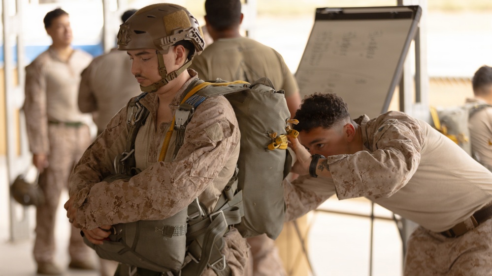 Two military personnel in desert camouflage uniforms are engaged in a parachute equipment inspection or fitting procedure. One soldier wearing a tactical helmet and parachute pack stands still while a second soldier closely inspects the parachute harness and pack from behind. Additional personnel and a whiteboard with writing are visible in the background.