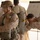 Two military personnel in desert camouflage uniforms are engaged in a parachute equipment inspection or fitting procedure. One soldier wearing a tactical helmet and parachute pack stands still while a second soldier closely inspects the parachute harness and pack from behind. Additional personnel and a whiteboard with writing are visible in the background.